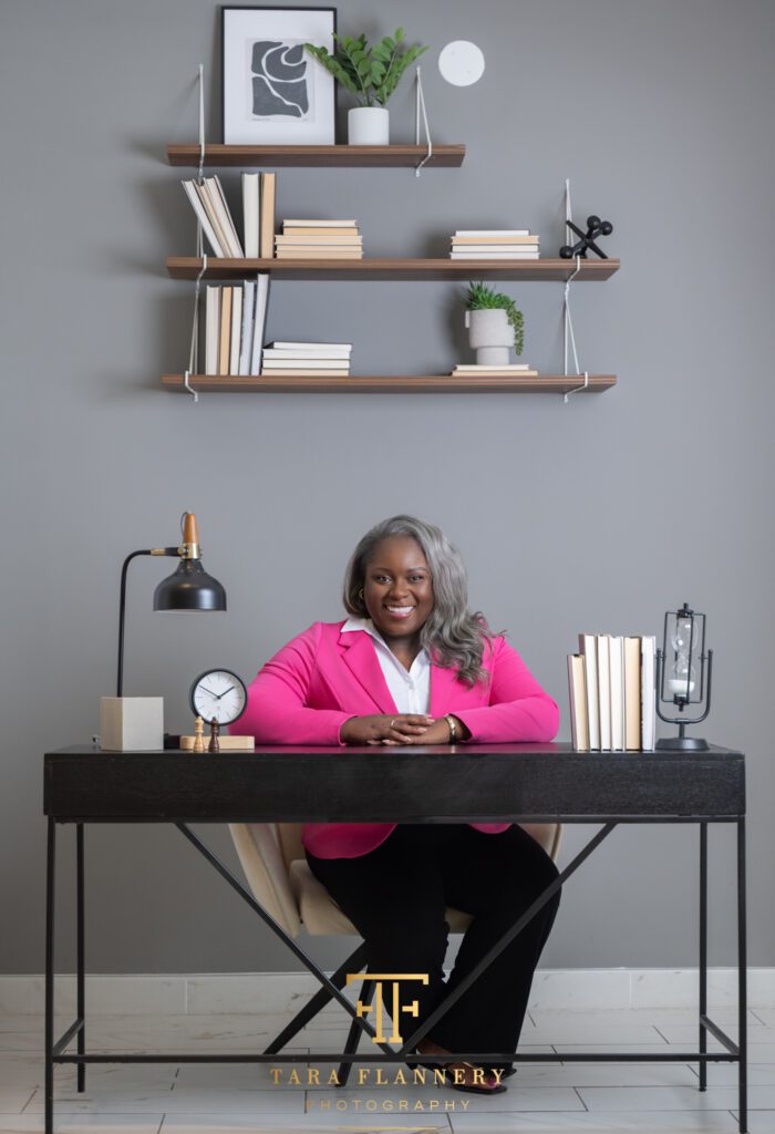 Professional business woman in pink jacket sits at a desk for a professional branding session.