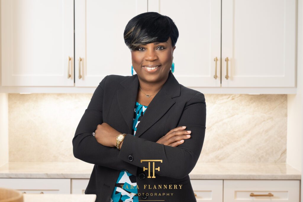 Woman in black suit jacket crossing her arms and smiling in a kitchen for a realtor headshot.